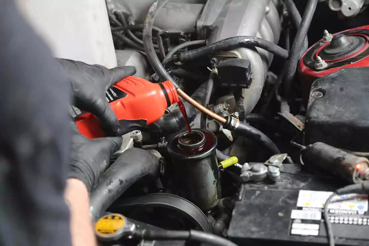 A mechanic wearing black nitrile gloves pours red power steering fluid into the reservoir.