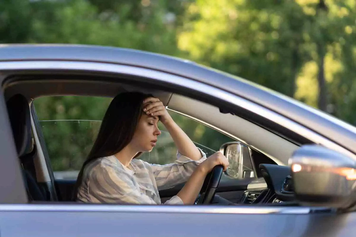 An unhappy female driver in a light blue car holds her forehead in despair.
