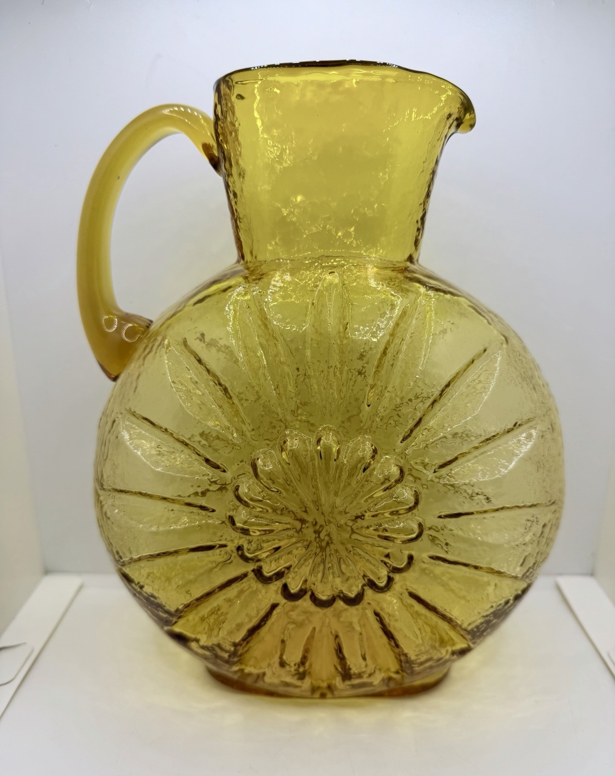 Amber glass pitcher vase with a sunburst sunflower design on a white background.