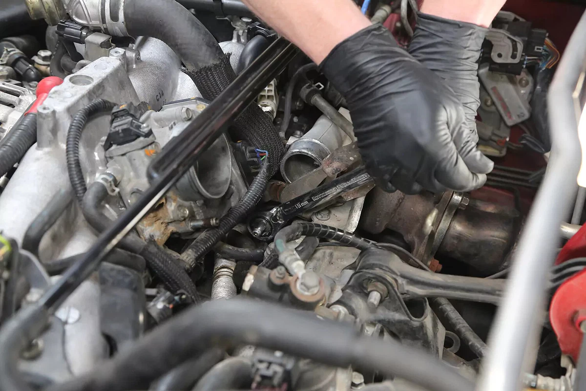 A mechanic wearing black gloves uses a ratchet to tighten a new PCV valve into place.