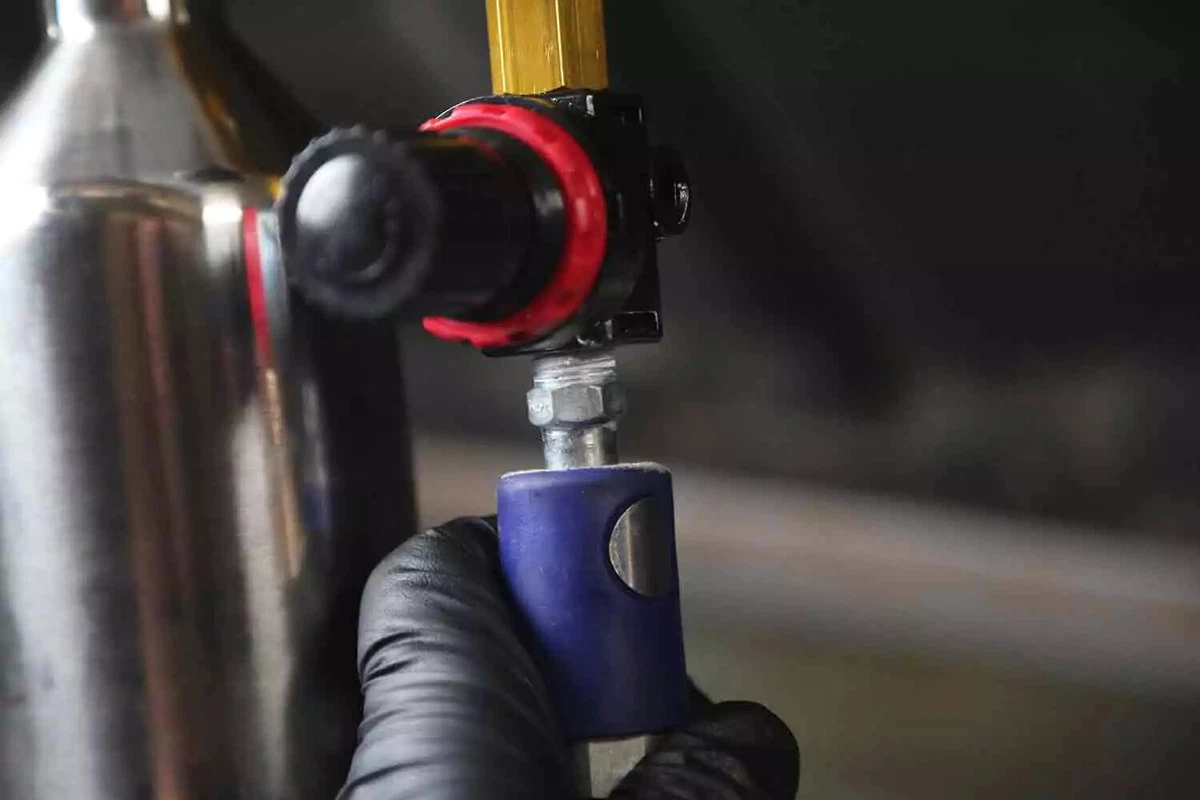 A mechanic wearing a black glove connects the line from an air compressor to the cleaning kit's regulator.