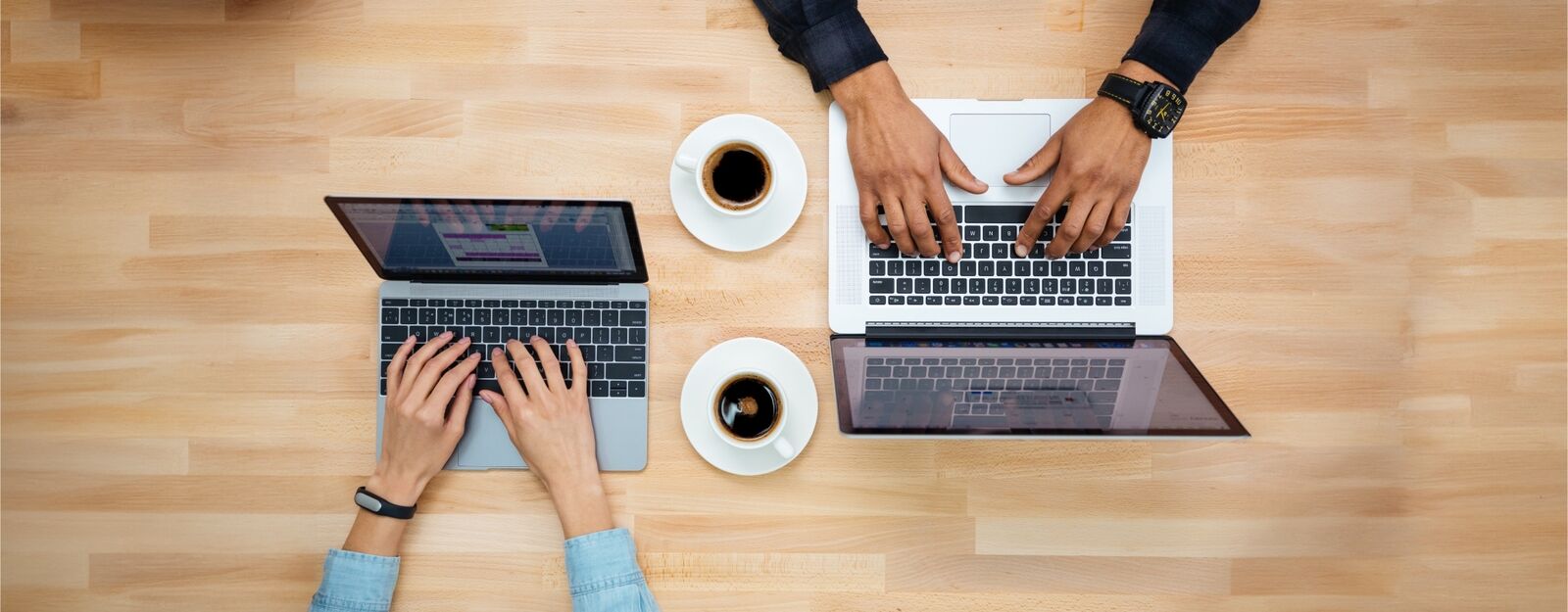 Zwei Menschen arbeiten parallel an Laptops, mit einer Tasse Kaffee dazwischen, Vogelperspektive.