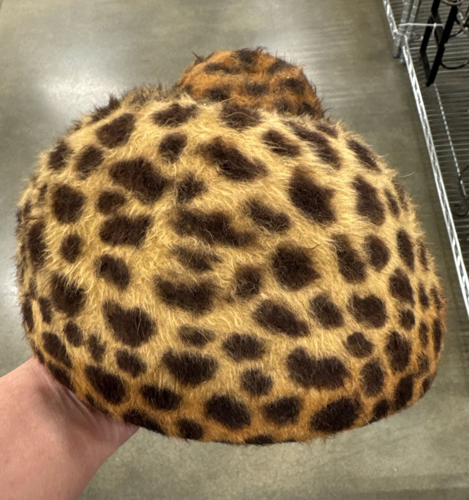 Leopard-print faux fur pillbox hat held up against a metal shelving unit, showing the domed crown and brim.