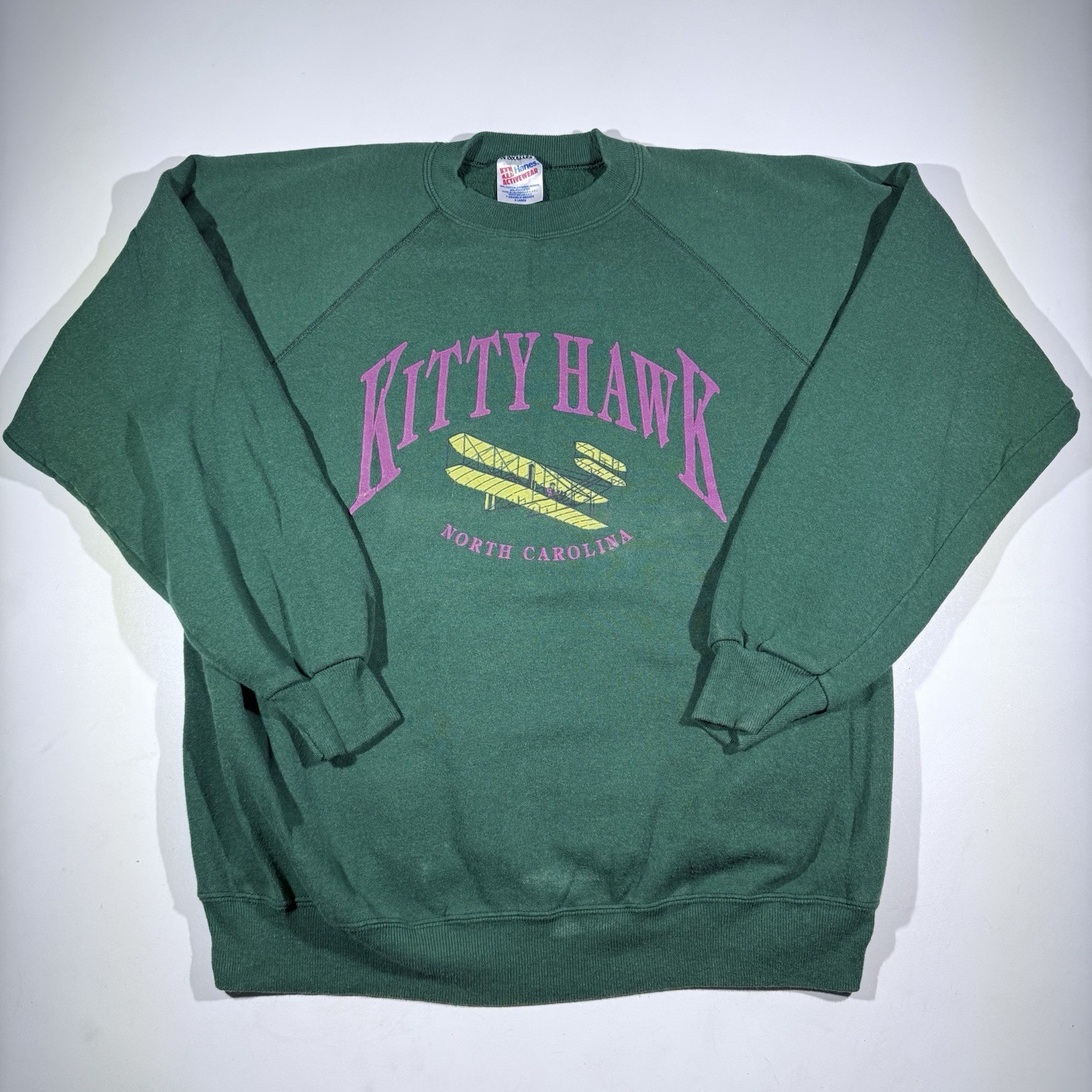 Green vintage Hanes Kitty Hawk sweatshirt with purple “Kitty Hawk North Carolina” text and surfboard graphic on front.