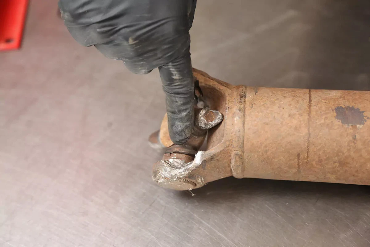 A mechanic wearing black gloves points to damage on a vehicle's shaft.