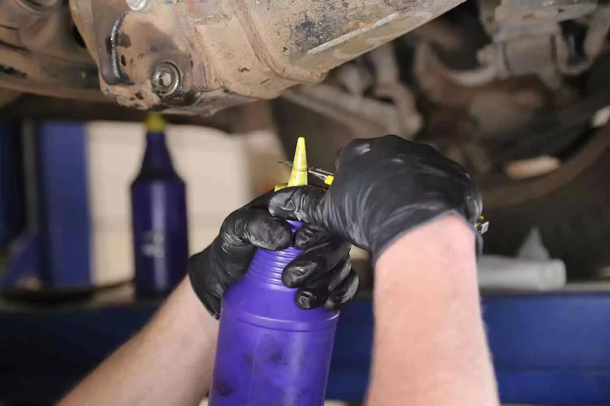 A mechanic wearing black gloves uses a knife to cut off the tip of a bottle of new differential fluid.