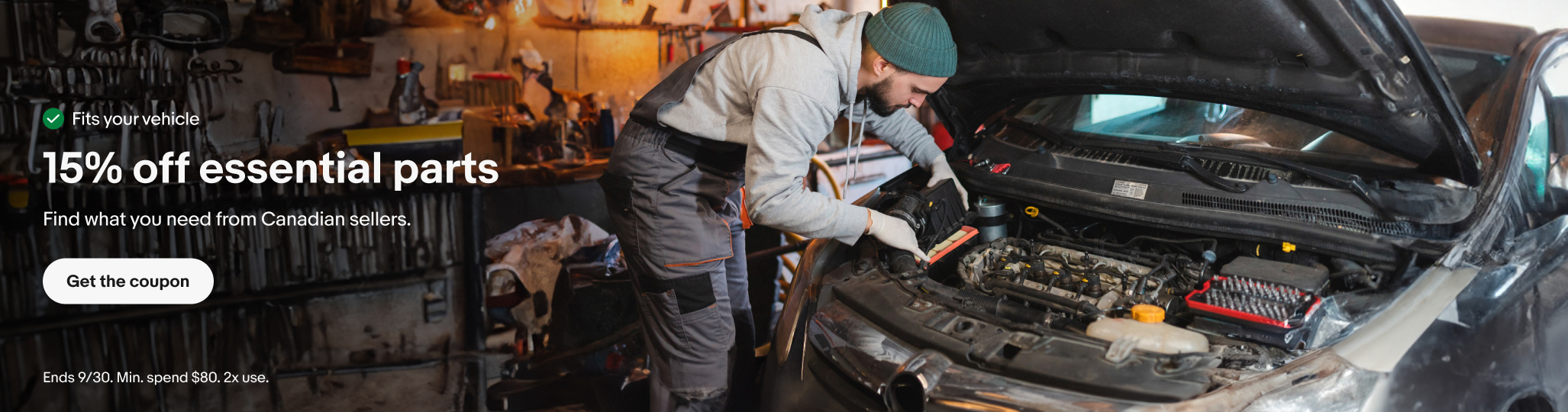 A man in a garage is working on a car engine. The hood of the car is open. The man is wearing a grey hoodie under dark overalls and a teal beanie.