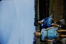 RED BORDER SLIDE Two girls in dresses outdoors, one sitting, one standing.