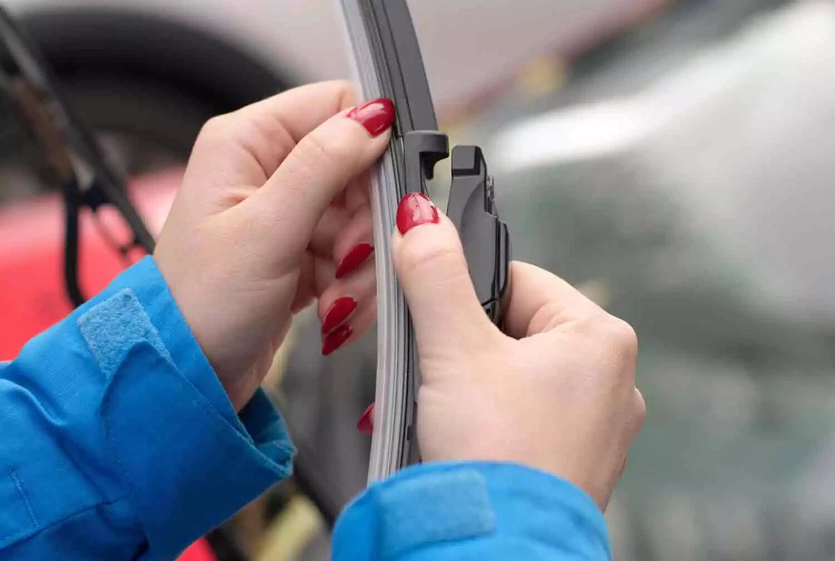 A driver wearing a teal jacket attaches a new windshield wiper blade to the wiper arm.