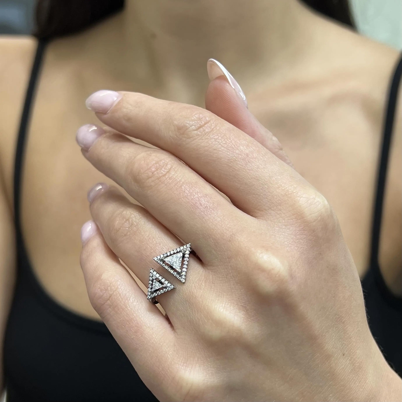 An image of a Messika diamond ring for women, showcased on a person's ring finger. The ring features a geometric design with multiple diamond-encrusted triangles. The photo is a close-up, with a focus on the hand and the ring at a slight angle, emphasizing the sparkle of the diamonds. The background is blurred, directing attention to the intricate details of the ring.