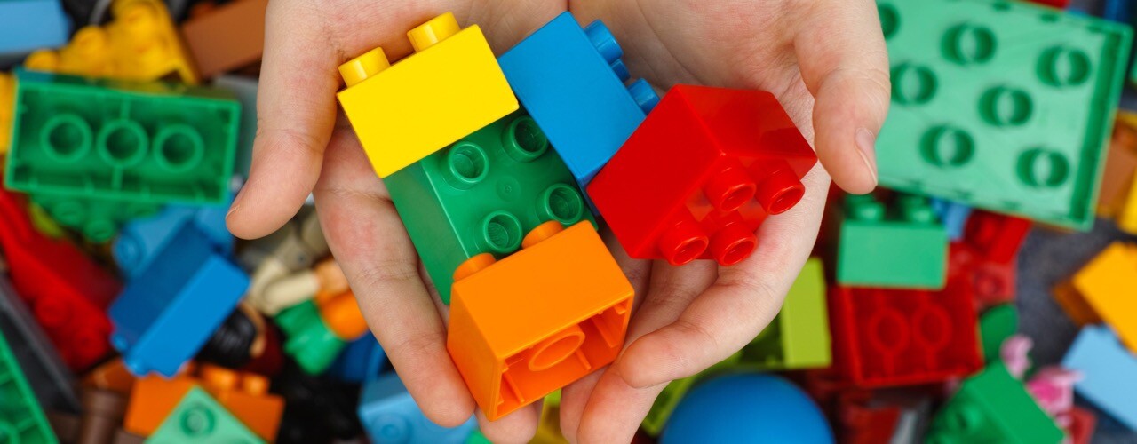 Aufnahme von zwei Händen, die fünf verschiedenfarbige DUPLO-Steine halten. Im Hintergrund sieht man noch viele weitere, bunte DUPLO-Steine.