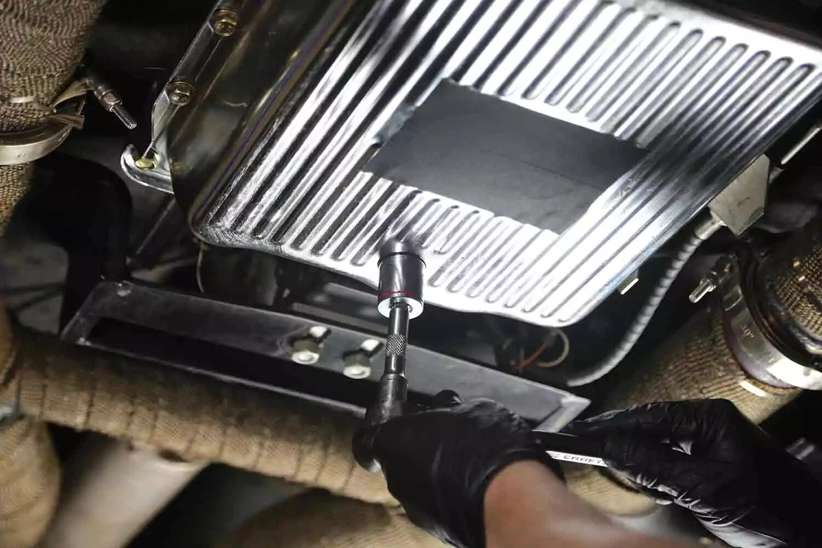 A mechanic wearing a black glove uses a socket and ratchet to loosen the drain plug of a vehicle's transmission pan.