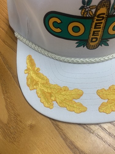 White vintage trucker hat with green brim, featuring yellow Coop Seed text and leaf graphics on the front panel.