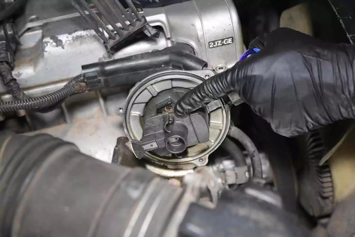 A mechanic wearing black gloves points to the rotor at the center of a vehicle's distributor cap.