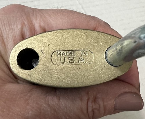Gold oval padlock with black keyhole, embossed text reading “MADE IN U.S.A.” on side, held by hand against white background.