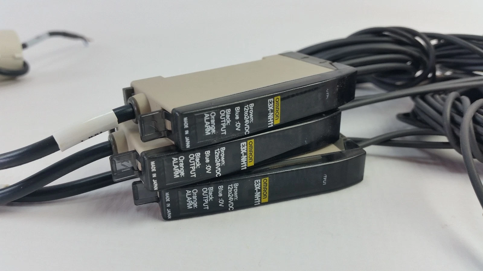 Stacked Omron E3X-NH11 amplifiers showing wiring label: brown 12–24 VDC, blue 0 V, black output, orange alarm