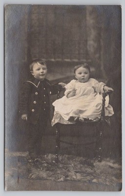 Zwei junge Geschwister, Junge im Matrosenanzug mit Baby auf Stuhl, RPPC um 1910