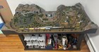 Z-scale model train layout with rocky cliffs, tunnel, pond, road intersection, and storage bins under the table.