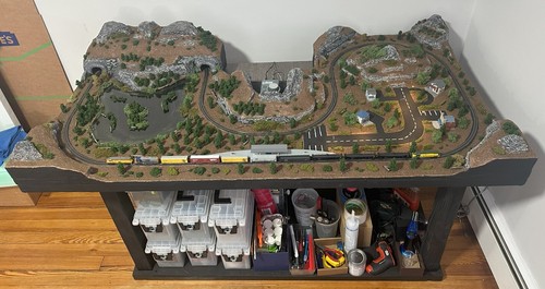 Z-scale model train layout with rocky cliffs, tunnel, pond, road intersection, and storage bins under the table.