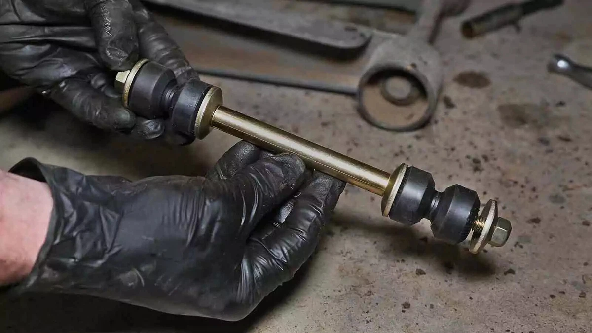 A mechanic wearing black nitrile gloves holds a new sway bar link, with a floor jack visible in the background.