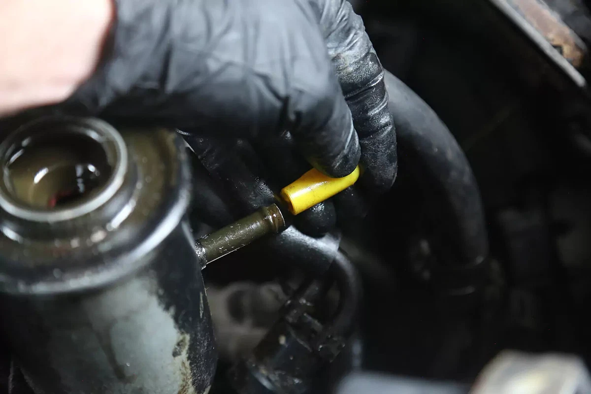 A mechanic wearing a black glove places a yellow rubber cap over the open power steering fluid return valve.