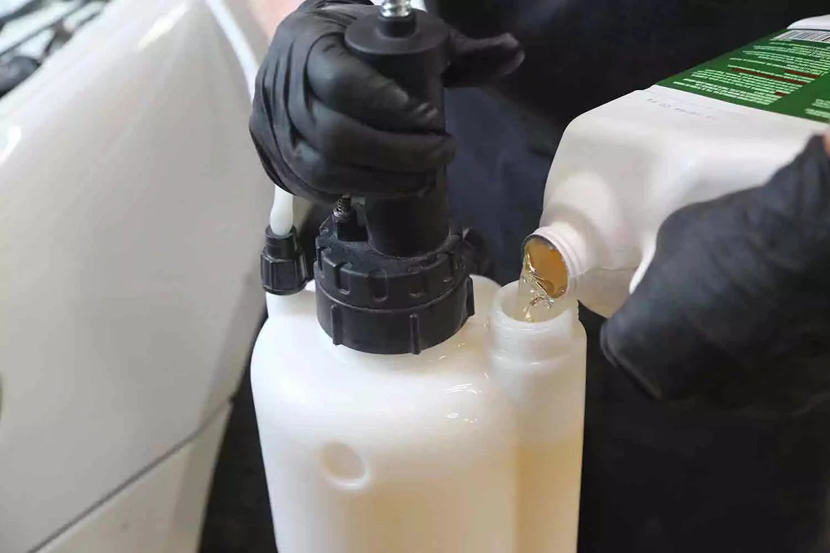 A mechanic wearing black gloves pours brake fluid from a white container into a vehicle's brake fluid reservoir.