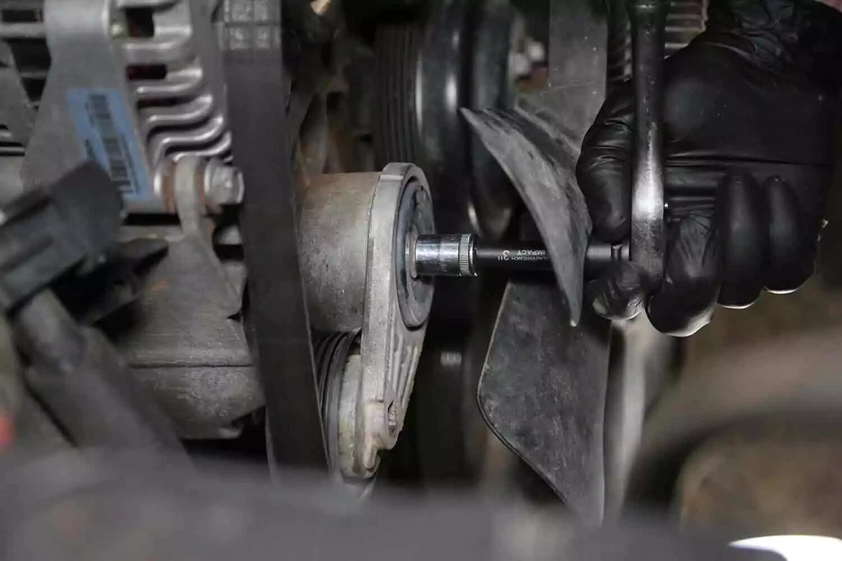 A mechanic wearing a black glove uses a ratchet to unbolt the serpentine belt tensioner from the engine block.