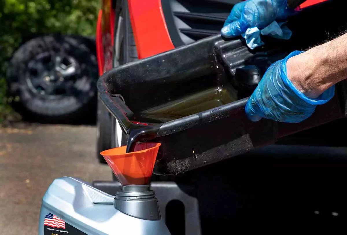 A backyard mechanic wearing blue nitrile gloves pours the dirty old oil into an empty five gallon oil container.