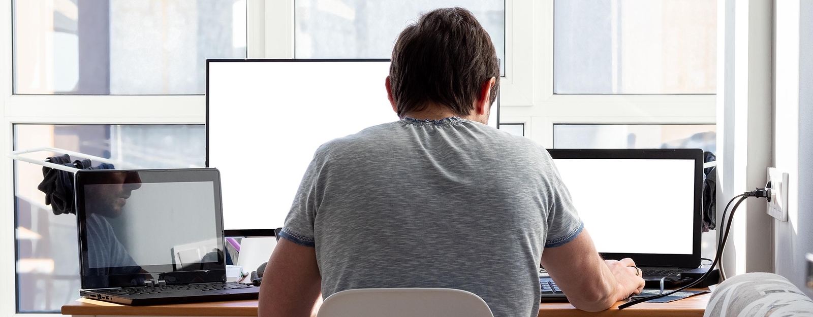 Ein junger Mann sitzt vor seinem Schreibtisch. Darauf stehen zwei Laptops und ein externer Bildschirm, an denen er arbeitet.