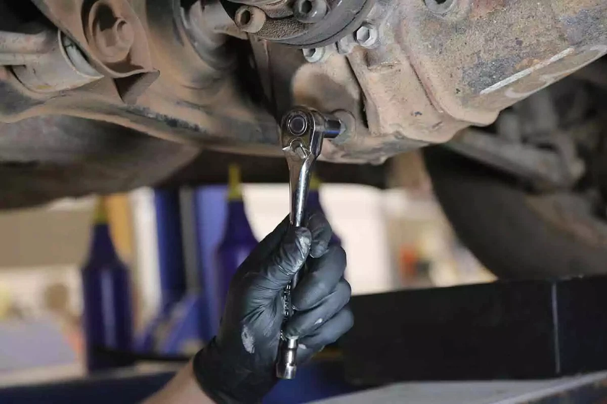 A mechanic wearing a black glove uses a socket to loosen the differential drain plug of a vehicle.