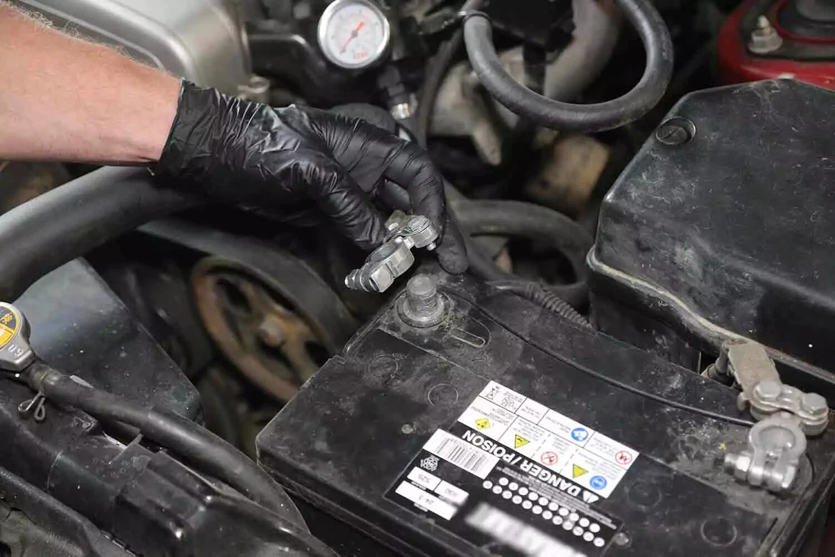 A mechanic wearing a black glove disconnects the negative terminal of a vehicle's battery.