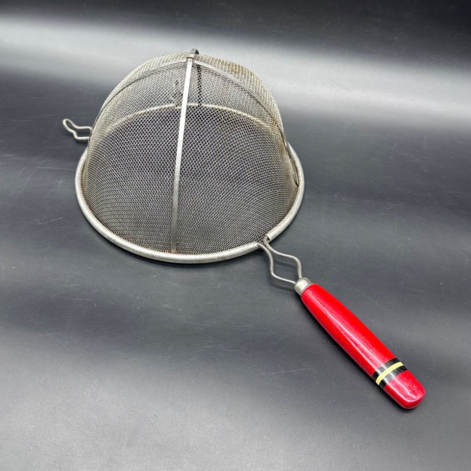 A gorgeous 1950s metal mesh strainer with a cherry-red turned wooden handle — the kind of functional sculpture that belongs on a kitchen peg board display.
