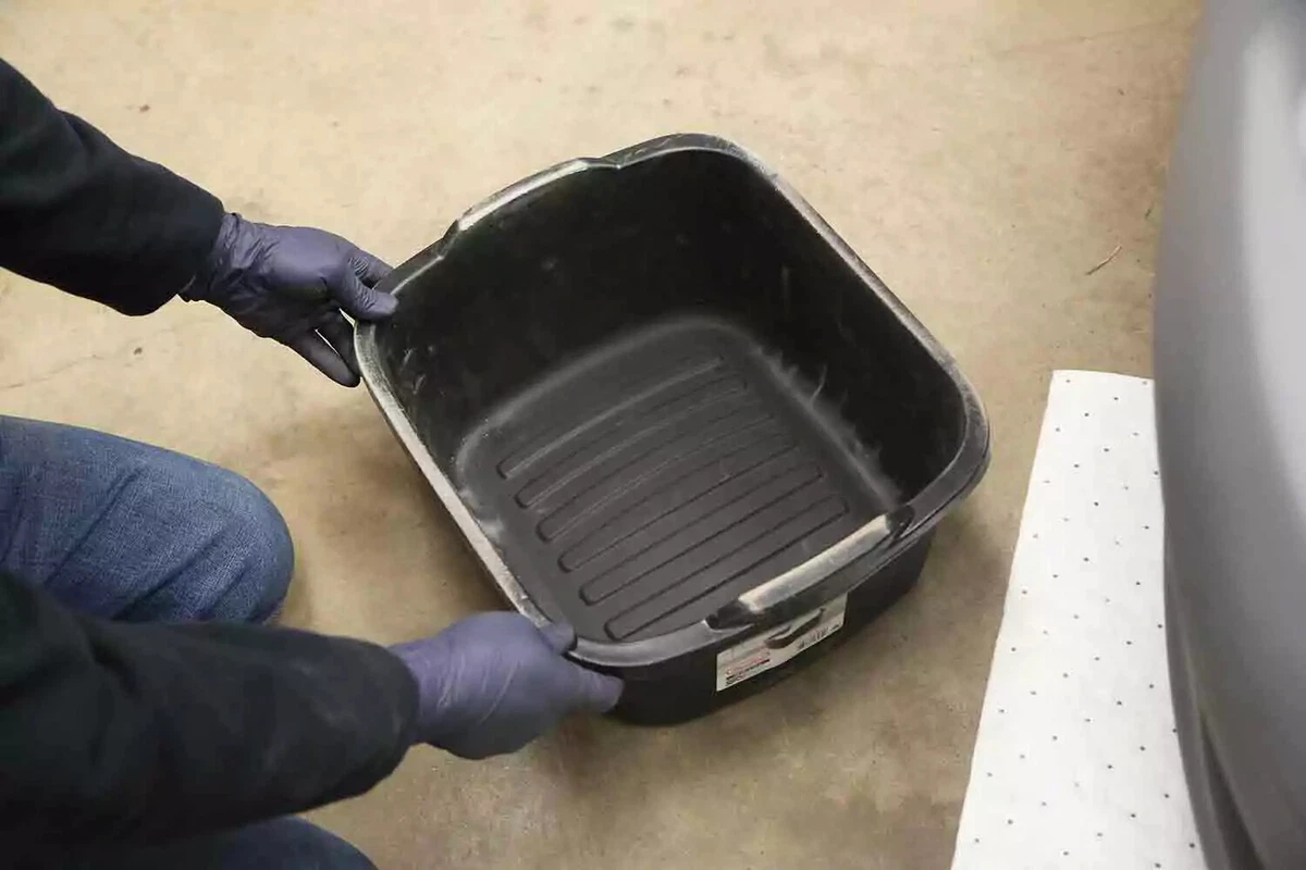 A mechanic wearing blue nitrile gloves slides a drain pan underneath a vehicle before flushing the engine coolant.