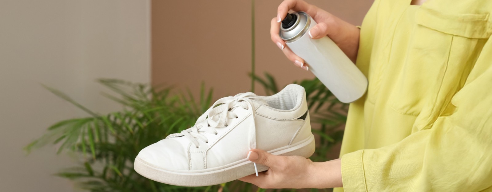 Eine Person in gelbem Oberteil hält einen weißen Sneaker in der Hand und besprüht ihn mit Imprägnierspray, im Hintergrund sind grüne Zimmerpflanzen zu sehen.