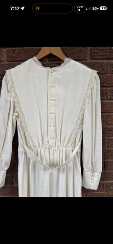 White vintage 70s midi prairie dress with lace trim, button front, and long sleeves on wooden hanger against brick wall.