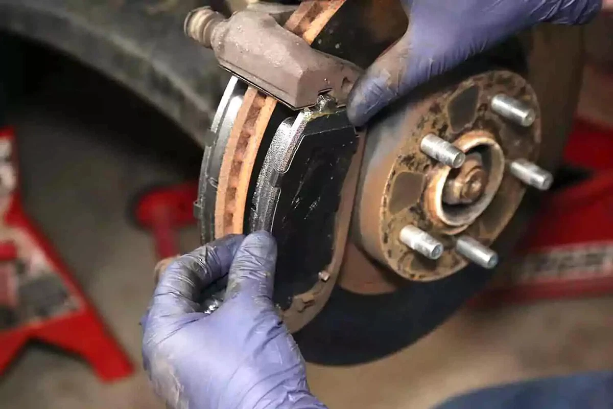 A mechanic wearing blue nitrile gloves loads a new brake pad into the brake caliper, with the vehicle supported with jack stands.