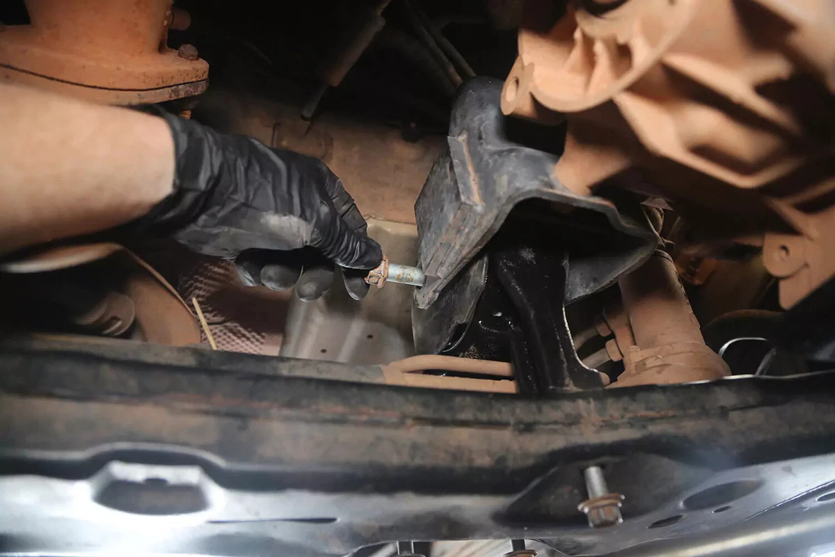 A mechanic wearing a black glove reinstalls a crossbolt into a transmission mount.