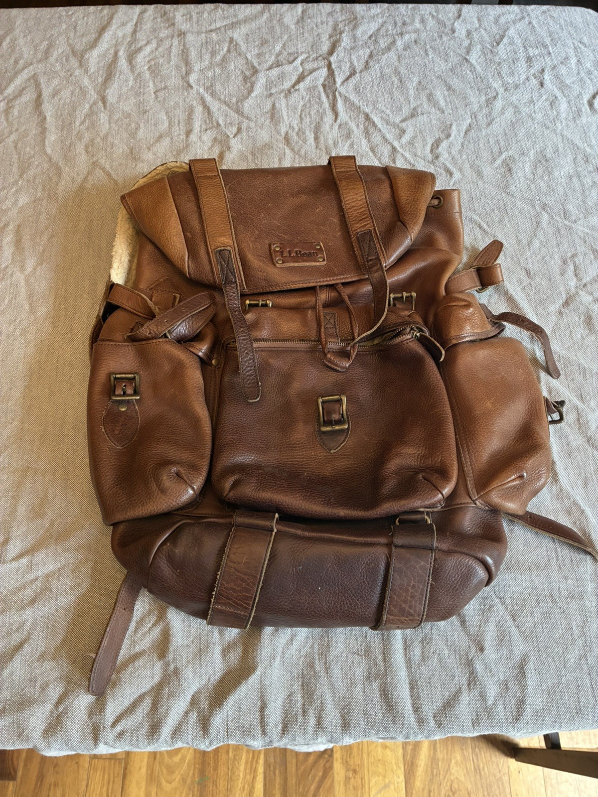 Worn brown leather L.L.Bean rucksack backpack with a flap top and multiple outer pockets, on a woven blanket.
