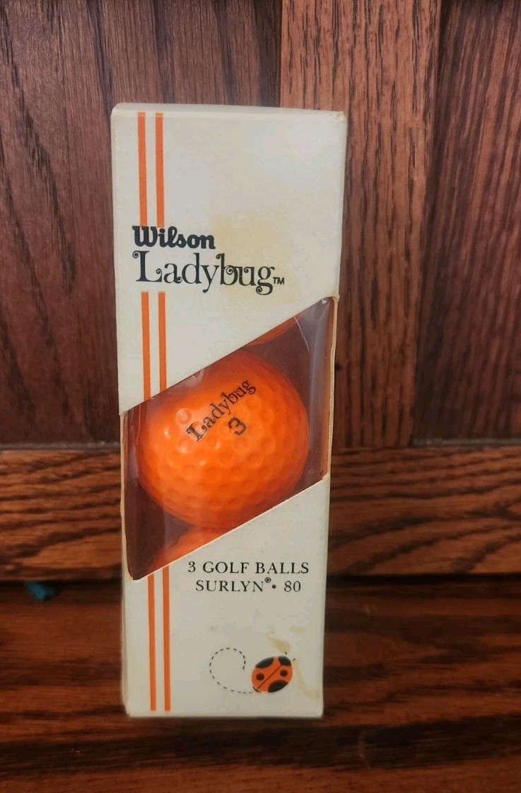 Three orange Wilson Ladybug golf balls in clear packaging, with white box labeled “3 Golf Balls Surlyn 80” and orange logo.