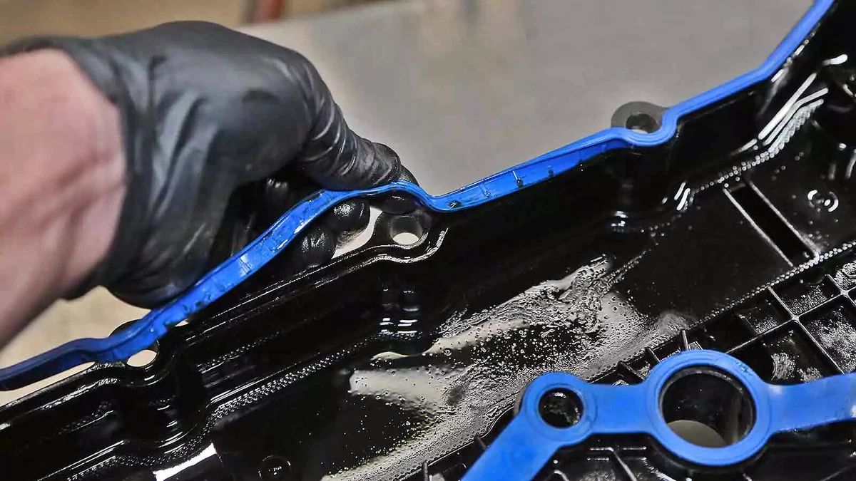 A mechanic wearing a black nitrile glove installs a fresh blue rubber gasket on a valve cover.