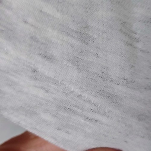 Close-up of worn white fabric with faint gray surface stain, small dark spot on left sleeve, and visible stitching.