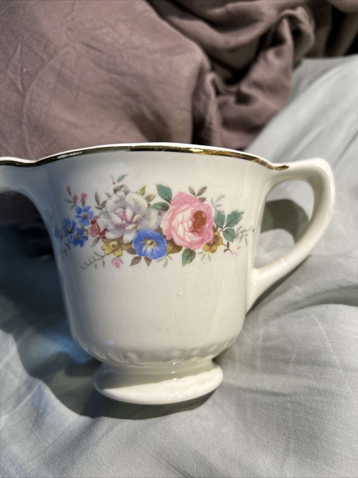 White ceramic mug with gold rim, pink rose floral print, and blue hydrangea accents on a gray fabric background.