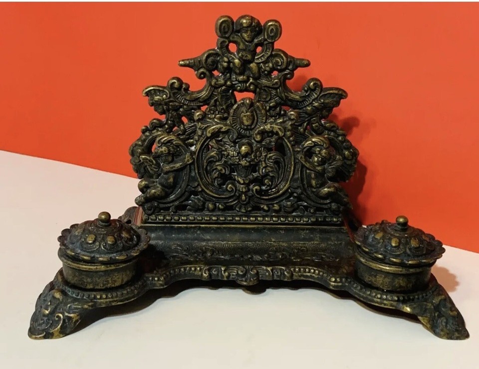 A brass inkwell featuring chubby Renaissance cherubs (putti) — absurdly ornate, visually delightful, and doubles as a letter holder.