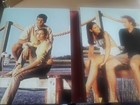 Three people in white clothing sitting on a wooden pier over a lake, with ropes and a sailboat in the background.