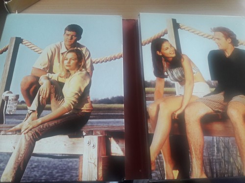 Three people in white clothing sitting on a wooden pier over a lake, with ropes and a sailboat in the background.
