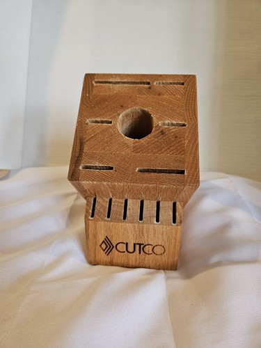 Rectangular wooden CUTCO knife storage block with 13 slots and a central circular hole, placed on a white fabric surface.