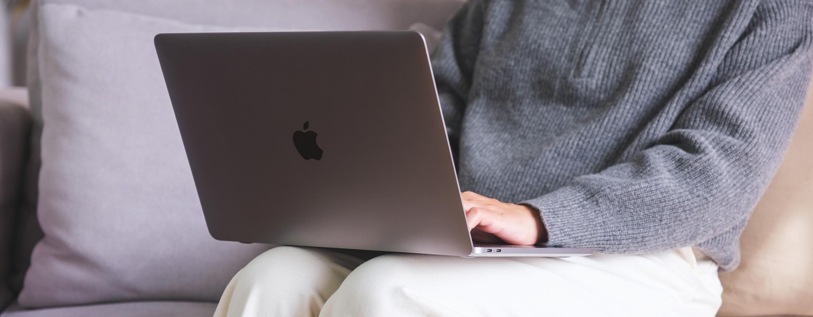Eine Person sitzt auf einem hellgrauen Sofa und hält ein aufgeklapptes MacBook auf dem Schoß. Kopf und Füße sind nicht zu sehen.