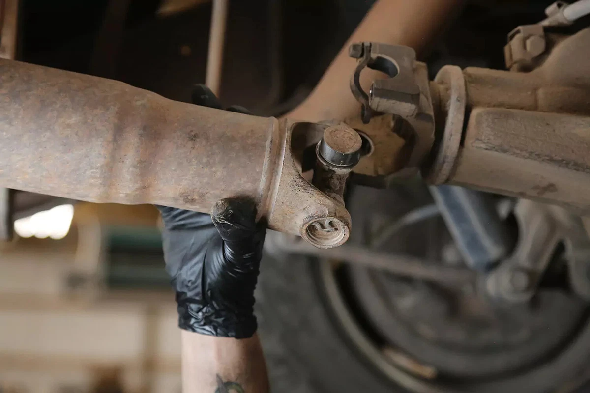 A mechanic wearing a black glove pushes a vehicle's driveshaft forward toward the transmission.