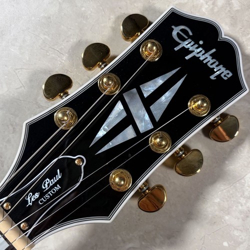Black Epiphone LP Custom electric guitar head with gold hardware, silver f-hole logo, and gold “Les Paul Custom” text.