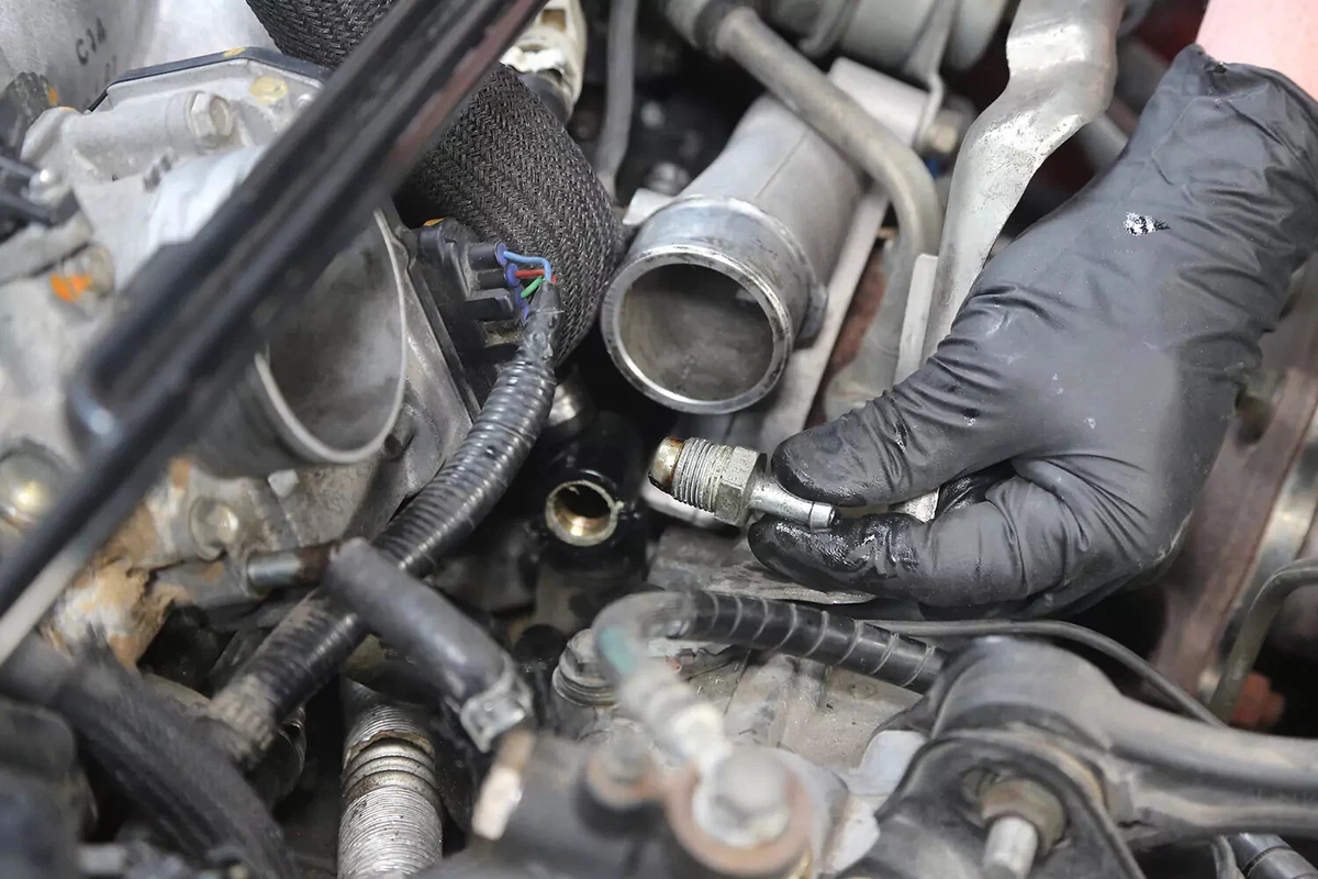 A mechanic wearing a black glove holds a vehicle's PCV valve after removing it.